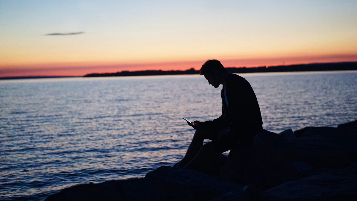 Man Sunset Mobile Rock Ocean River Мужчина сидит на камнях на берегу моря океана и смотрит в телефон на фоне заката солнца