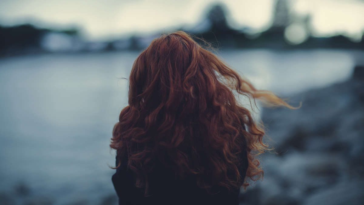 Woman Red-haired Curls Wind Landscape Blur BG Девушка рыжеволосая стоит на фоне размытого пейзажа гор