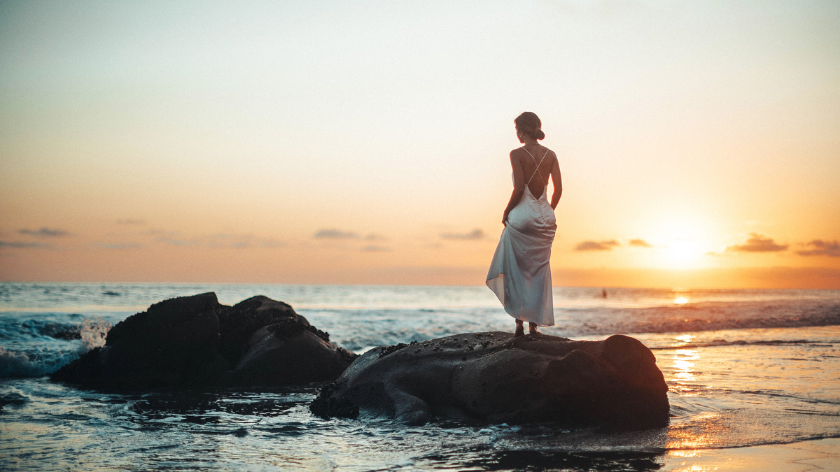 Woman Blonde Dress Sunset Rocks Ocean Девушка блондинка в платье стоит на камнях на берегу океана на фоне заката солнца