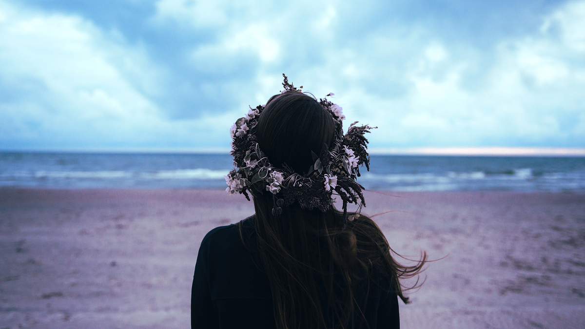 Woman Blonde Ocean Flowers Hair Beach Девушка брюнетка на фоне прибоя океана в холодный пасмурный день с венком из цветов на голове на пляже